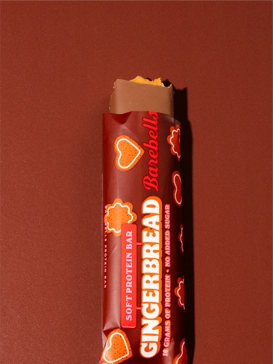 A Barebella Gingerbread soft protein bar wrapped in a dark red wrapper with gingerbread cookie illustrations, shown on a solid dark reddish-brown background with a bite taken from the top.