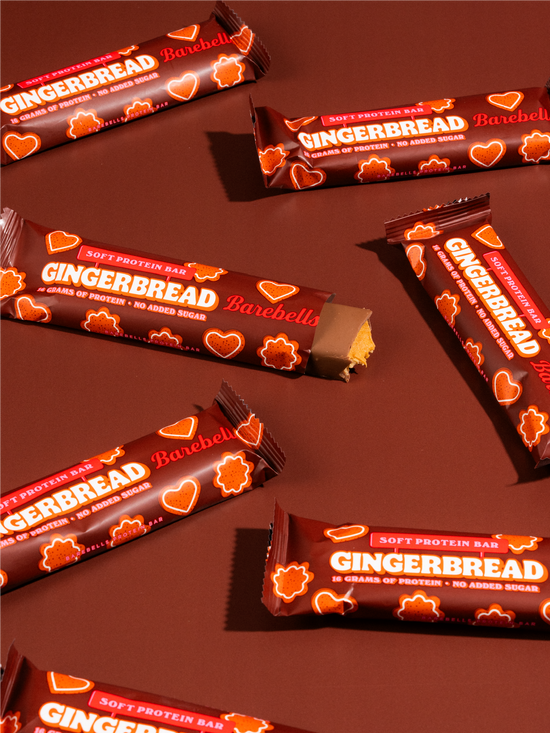 Several Barebella Gingerbread soft protein bars are arranged on a dark brown background. The packaging is dark brown with red and white text and orange heart-shaped cookie illustrations.