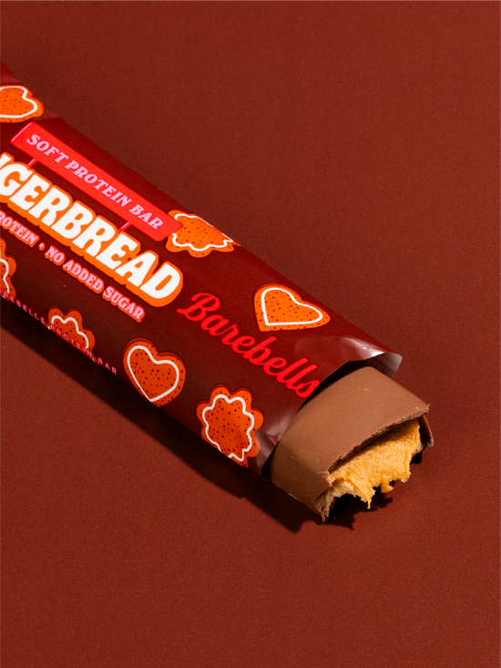 A packaged Barebells Gingerbread Soft Protein Bar, partially unwrapped to show the chocolate-coated cookie and cream filling, set against a solid dark reddish-brown background.